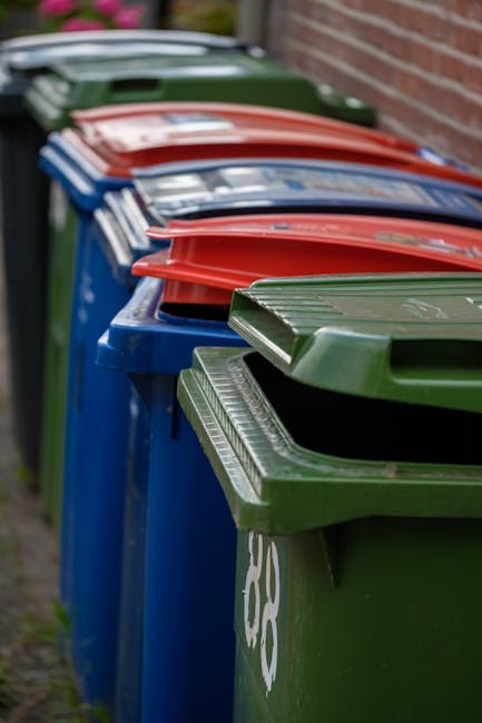 A row of residential wheelie bins lined up along a pavement beside a brick wall, with each bin featuring a large, hinged lid. The bins are primarily bright blue, green, and red in colour, with some displaying white recycling symbols or markings. The green bin closest to the camera has a textured lid with ridges, and the blue bins behind have smooth surfaces. The scene suggests a typical waste disposal area outside a household or administrative building, with the bins arranged neatly for waste collection. The background shows a slight blur of greenery and part of a building, indicating an outdoor urban or suburban setting. The lighting is natural, with soft daylight illuminating the scene, emphasizing the colours and textures of the plastic bins. This visual relates to private waste collection services, such as those provided by Rubbish Clearance Catford, illustrating the organised approach to rubbish removal at residential properties.