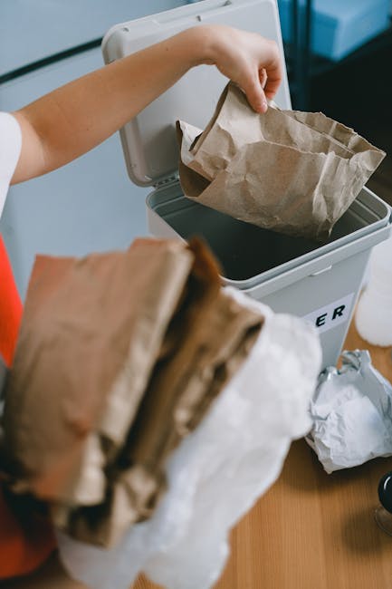 A person is shown discarding a brown paper bag into a closed, grey rubbish bin with a black interior, positioned on a wooden surface. In the foreground, another crumpled paper bag and some waste material are being held, indicating ongoing rubbish disposal. The bin appears to be part of a waste collection service, possibly representing private waste handling or alternative rubbish removal options. The background includes a glimpse of a light blue wall and a white surface, suggesting an indoor setting such as an office or kitchen area. The scene emphasizes the process of rubbish disposal, with the focus on the open bin and the act of placing waste inside, aligning with waste clearance practices that may differ from council-led collection. The lighting is natural or softly diffused, providing clear visibility of the materials and textures, such as the roughness of the paper bags and the smooth plastic of the bin, supporting a professional and factual presentation related to rubbish removal services provided by Rubbish Clearance Catford.