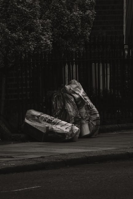 A street scene featuring several large, crumpled black rubbish bags placed along the pavement curbside, with some bags leaning against a dark metal fence and others positioned directly on the asphalt. The bags appear to be made of thick, glossy plastic material, with visible creases and folds. Behind the fence, dense foliage from trees or bushes is partially visible, and the background includes a building structure with a dark, patterned exterior. The overall atmosphere is subdued, with muted lighting suggesting overcast conditions or low natural light levels. This scene illustrates typical private waste disposal or rubbish collection during street cleaning or rubbish removal services, possibly undertaken by a company such as Rubbish Clearance Catford, in alignment with the theme of flat rubbish removal and alternative waste handling methods.