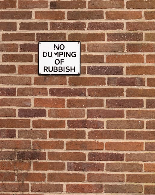 A rectangular metal sign mounted on a red brick wall displaying the words 'NO DUMPING OF RUBBISH' in black uppercase letters, with the word 'DUMPING' partially obscured by a perceived hole or tear in the sign. The brick wall behind the sign consists of evenly spaced bricks with a reddish-brown hue, and visible mortar joints. The scene appears to be outdoors, with natural lighting casting slight shadows on the wall, and the sign's purpose indicating a restricted area for waste disposal in alignment with private rubbish removal services such as those offered by Rubbish Clearance Catford.