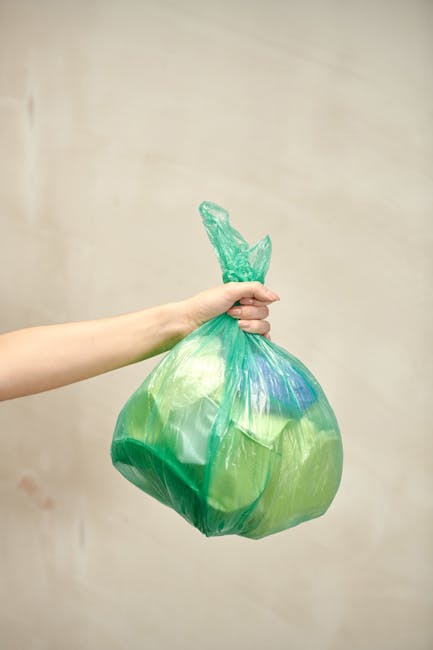 A street scene featuring several large, crumpled black rubbish bags placed along the pavement curbside, with some bags leaning against a dark metal fence and others positioned directly on the asphalt. The bags appear to be made of thick, glossy plastic material, with visible creases and folds. Behind the fence, dense foliage from trees or bushes is partially visible, and the background includes a building structure with a dark, patterned exterior. The overall atmosphere is subdued, with muted lighting suggesting overcast conditions or low natural light levels. This scene illustrates typical private waste disposal or rubbish collection during street cleaning or rubbish removal services, possibly undertaken by a company such as Rubbish Clearance Catford, in alignment with the theme of flat rubbish removal and alternative waste handling methods.