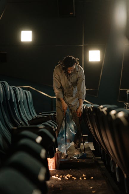 A person with dark, curly hair tied back and wearing a grey jumpsuit is seen collecting scattered rubbish from the floor of an auditorium or cinema. They are bent over, holding a large clear plastic bag to gather the debris, which includes crumpled paper and small discarded items. The individual is positioned between rows of dark teal or green plastic seats that are attached to the floor, with their backs facing the camera. The room is dimly lit, with two bright square lights mounted on the dark wall behind, creating a stark contrast and illuminating the scene subtly. The environment appears to be an indoor venue, possibly a screening room or theatre, with a dark, modern decor. The scene conveys an act of cleaning or clearing waste, aligned with independent rubbish removal or on-site clearance services. Natural textures of the seats and the scattered litter, along with the utilitarian setting, support the context of waste collection or disposal, as managed by a private rubbish clearance company like Rubbish Clearance Catford.