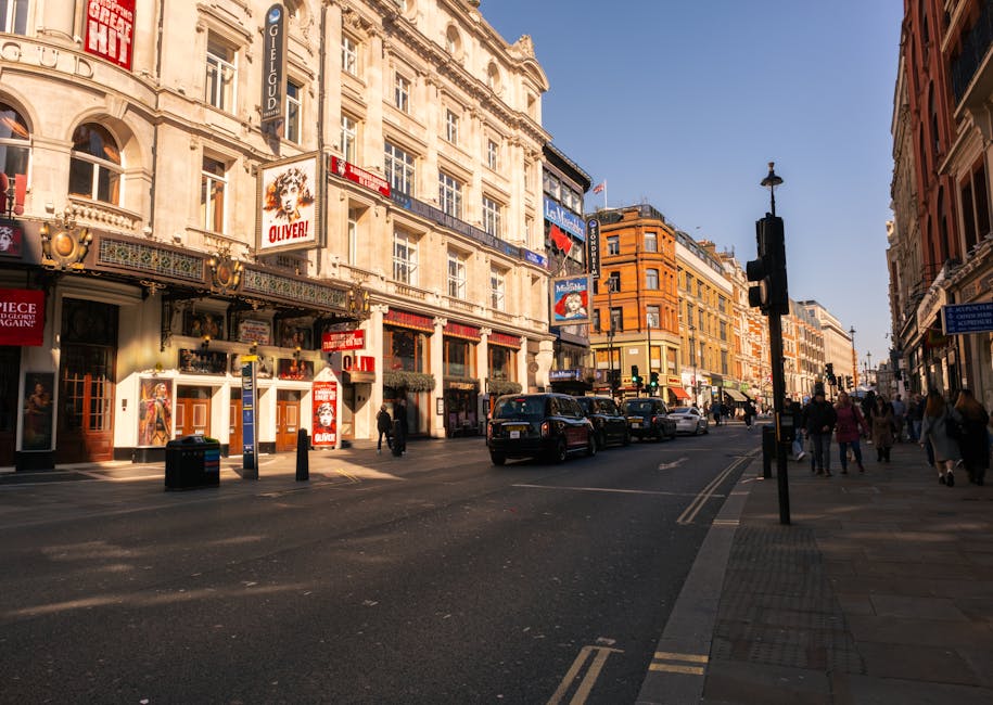 The image depicts a lively urban street scene during daylight hours, featuring historic buildings with ornate facades and large windows lining the street. The buildings are primarily off-white with decorative detailing, contrasting with the adjacent structure, which is modern and constructed from red-brick materials. Signage for theatres and entertainment venues, including posters and illuminated signs, adorn the fronts of several buildings, suggesting a commercial entertainment district. The street is populated with pedestrians walking along the wide sidewalk, some crossing at the pedestrian crossings, while others are gathered near shop entrances. Several vehicles, including a black taxi and small cars, are parked along the curb or moving through the street, which is marked with double yellow lines indicating parking restrictions. A tall black traffic light stands prominently near the intersection, and a street lamp is visible further down the road. The scene is bright, with clear blue sky overhead, and the warm sunlight casts shadows on the buildings and pavement, creating a vibrant yet orderly city atmosphere typical of a busy commercial district within a UK town or city. This setting can be associated with urban rubbish removal and private waste handling services, such as those offered by Rubbish Clearance Catford, who manage waste collection from such active city environments.