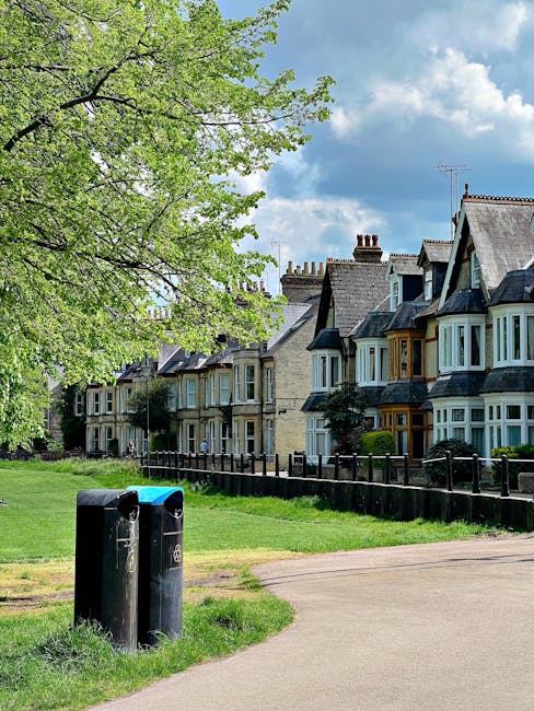 A row of traditional terraced houses with bay windows and pitched roofs, situated along a tree-lined street in a residential area. The houses are constructed from brick and stone, with some featuring painted wood detailing around the windows. In the foreground, two black public waste bins are positioned on a paved footpath, with a small grassy verge separating them from the pavement. To the left, a mature tree with fresh green leaves extends its branches over the scene, creating a canopy that partially shades the area. The background shows a partly cloudy sky with patches of blue, indicating a bright day. The setting appears calm and well-maintained, with a low wooden fence running alongside the houses, separating the front gardens from the pavement. This scene relates to local urban waste collection points within a residential neighborhood, emphasizing the importance of proper rubbish disposal and waste management practices often supported by independent rubbish clearance services like Rubbish Clearance Catford.