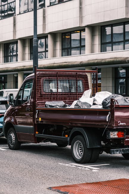 A small burgundy flatbed truck parking on a city street in front of a modern office building with large glass windows and a concrete facade. The vehicle's open cargo area is filled with various types of waste, including plastic bags, cardboard boxes, and loose debris, indicating recent rubbish collection or clearance. The truck features a protective metal grid behind the cab, which is visible through the rear window. Surrounding the truck, there are parked cars and a lamppost positioned adjacent to the vehicle. The overall scene suggests an urban environment where on-site rubbish removal or private waste handling services are taking place, aligning with independent clearance options typically offered by companies like Rubbish Clearance Catford.