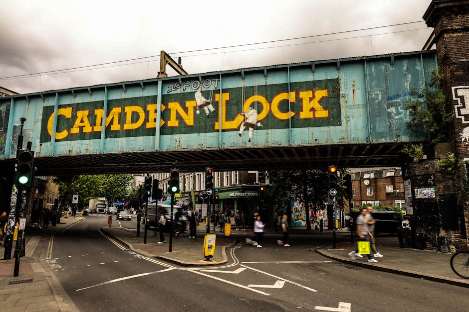 A large, weathered green railway bridge with rusted metal surfaces spans over a busy urban street, prominently displaying the painted signage 'CAMDEN LOCK' in bold yellow letters. Two playful illustrations of children hanging from the letters add visual interest to the sign. Beneath the bridge, multiple traffic lights and pedestrian crossings guide vehicle and pedestrian flow, with some lights illuminated in green and red. The street area features a range of moving vehicles, including cars and small trucks, along with pedestrians walking on the sidewalks. Some individuals are crossing the street, while others stand at bus stops or near shops visible in the background, which include brick buildings and storefronts decorated with graffiti and street art. The environment conveys a lively cityscape typical of Camden, with overcast skies casting diffuse natural light over the scene. The structure of the bridge, with its horizontal beams and supporting pillars, emphasizes the urban infrastructure, supporting local transit and potentially serving as an element of private or independent rubbish removal activities in busy areas like this, consistent with services offered by Rubbish Clearance Catford.